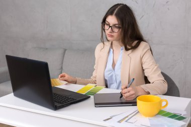 Masada oturan genç bir tasarımcı kız tablete resim çizer. Bilgisayarda iş hesaplamaları yapar.
