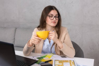 Masada oturan genç bir tasarımcı kız tablete resim çizer. Bilgisayarda iş hesaplamaları yapıyor. Bir kupa kahveden içmek.