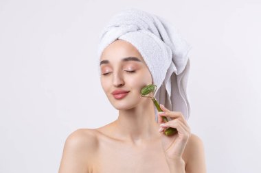 Kalın kaşlı güzel bir kız, beyaz arka planda mükemmel bir ten, kafasında havlu, güzel bir fotoğraf. Kozmetik rulo tutuyor..