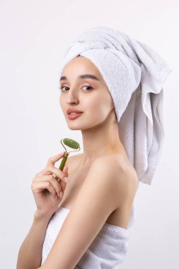 Kalın kaşlı güzel bir kız, beyaz arka planda mükemmel bir ten, kafasında havlu, güzel bir fotoğraf. Kozmetik rulo tutuyor..