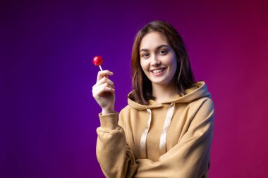 Kapüşonlu bir kadının portresi, stüdyoda mor renkli arka planda güzel makyajlı kırmızı yuvarlak bir lolipopu yalayan kızlar..