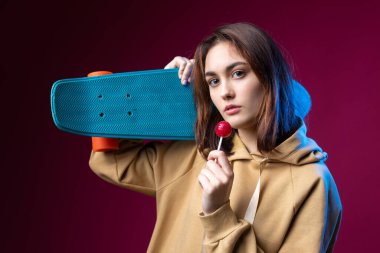 Kapüşonlu güzel, modaya uygun bir hippi kız elinde mor bir stüdyo geçmişiyle bir kaykay tutuyor. Lolipop yiyor..