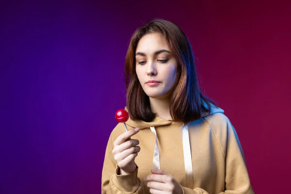 Portrait of a woman in a hoodie, girls licking a red round lollipop with beautiful makeup on a purple trendy background in the studio.