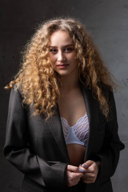 Stüdyoda uzun saçlı, ceketli, kızıl saçlı, kıvırcık bir kızın portresi. Kıvrımlı göğüslerini sütyen içinde gösteriyor..