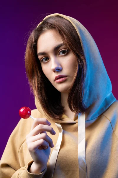 Portrait of a woman in a hoodie, girls licking a red round lollipop with beautiful makeup on a purple trendy background in the studio.