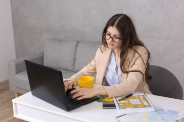 Masada oturan genç bir tasarımcı kız tablete resim çizer. Bilgisayarda iş hesaplamaları yapar.