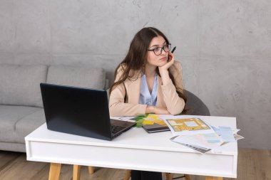 Masada oturan genç bir tasarımcı kız tablete resim çizer. Bilgisayarda iş hesaplamaları yapar.