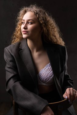 Stüdyoda uzun saçlı, ceketli, kızıl saçlı, kıvırcık bir kızın portresi. Kıvrımlı göğüslerini sütyen içinde gösteriyor..