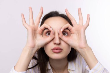 Beyaz stüdyo arka planında güzel bir jest sergileyen ve elleriyle dürbün yapan çekici genç kadının portresini kapat..