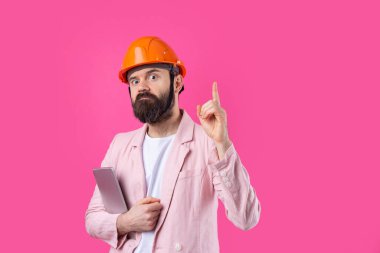 Genç inşaat mühendisinin portresi kırmızı stüdyo arka planında duran pembe ceketli turuncu bir şapka giyer. Elinde tablet olan bir adam..