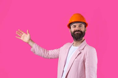 Genç inşaat mühendisinin portresi kırmızı stüdyo arka planında duran pembe ceketli turuncu bir şapka giyer. Adam eliyle işaret ediyor..