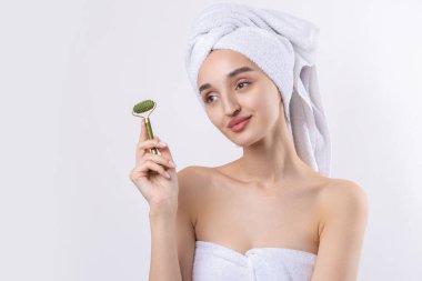Kalın kaşlı güzel bir kız, beyaz arka planda mükemmel bir ten, kafasında havlu, güzel bir fotoğraf. Kozmetik rulo tutuyor..