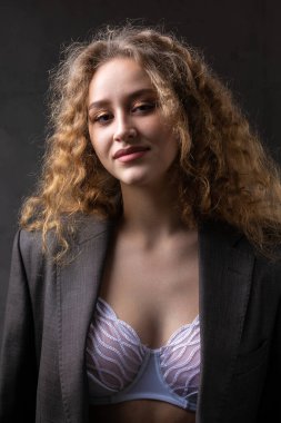Stüdyoda uzun saçlı, ceketli, kızıl saçlı, kıvırcık bir kızın portresi. Kıvrımlı göğüslerini sütyen içinde gösteriyor..