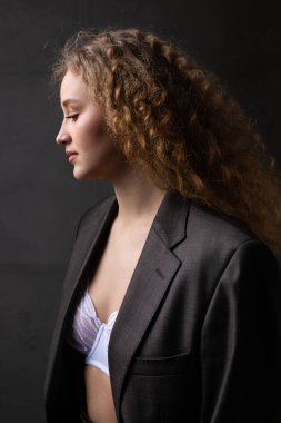 Stüdyoda uzun saçlı, ceketli, kızıl saçlı, kıvırcık bir kızın portresi. Kıvrımlı göğüslerini sütyen içinde gösteriyor..