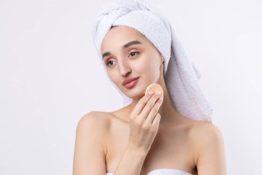 Kalın kaşlı güzel bir kız, beyaz arka planda mükemmel bir ten, kafasında havlu, güzel bir fotoğraf. Kozmetik sünger tutuyor..