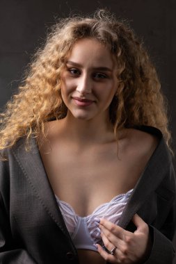 Stüdyoda uzun saçlı, ceketli, kızıl saçlı, kıvırcık bir kızın portresi. Kıvrımlı göğüslerini sütyen içinde gösteriyor..