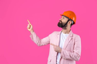Genç inşaat mühendisinin portresi kırmızı stüdyo arka planında duran pembe ceketli turuncu bir şapka giyer. Adam eliyle işaret ediyor..
