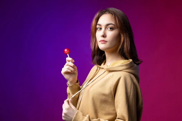 女の肖像画でAパーカー 女の子なめるA赤ラウンドロリポップとともに美しいメイク上の紫のトレンディーな背景でザ スタジオ — ストック写真