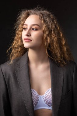 Stüdyoda uzun saçlı, ceketli, kızıl saçlı, kıvırcık bir kızın portresi. Kıvrımlı göğüslerini sütyen içinde gösteriyor..
