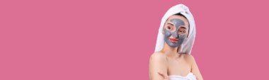 Pasifiğin pembe arka planında beyaz havluyla gri kozmetik maskeli bir kadın..