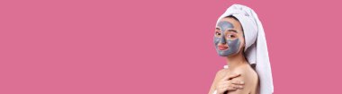 Pasifiğin pembe arka planında beyaz havluyla gri kozmetik maskeli bir kadın..