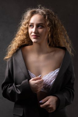 Stüdyoda uzun saçlı, ceketli, kızıl saçlı, kıvırcık bir kızın portresi. Kıvrımlı göğüslerini sütyen içinde gösteriyor..