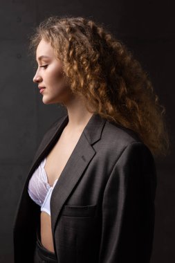 Stüdyoda uzun saçlı, ceketli, kızıl saçlı, kıvırcık bir kızın portresi. Kıvrımlı göğüslerini sütyen içinde gösteriyor..