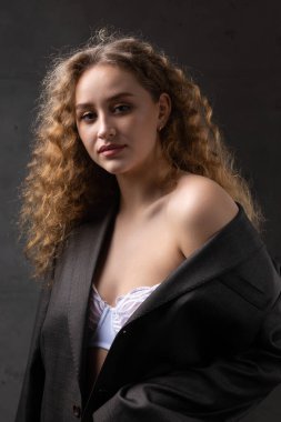 Stüdyoda uzun saçlı, ceketli, kızıl saçlı, kıvırcık bir kızın portresi. Kıvrımlı göğüslerini sütyen içinde gösteriyor..