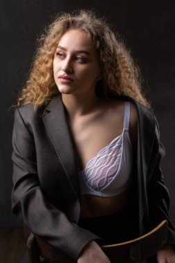 Stüdyoda uzun saçlı, ceketli, kızıl saçlı, kıvırcık bir kızın portresi. Kıvrımlı göğüslerini sütyen içinde gösteriyor..
