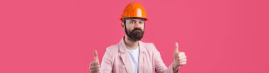 Genç inşaat mühendisinin portresi kırmızı stüdyo arka planında duran pembe ceketli turuncu bir şapka giyer. Adam eliyle işaret ediyor..