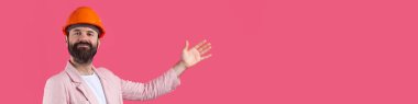 Genç inşaat mühendisinin portresi kırmızı stüdyo arka planında duran pembe ceketli turuncu bir şapka giyer. Adam eliyle işaret ediyor..