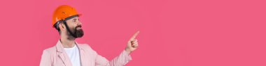 Genç inşaat mühendisinin portresi kırmızı stüdyo arka planında duran pembe ceketli turuncu bir şapka giyer. Adam eliyle işaret ediyor..