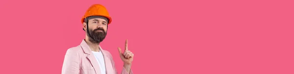年轻的建筑工程师头戴橙色硬礼帽 穿着粉色夹克 站在红色工作室的背景 那个人用手指指 — 图库照片