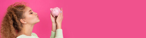 A cute young girl with curly red hair holds a piggy bank, a pink piglet in her hands. The concept of wealth and safety of money.