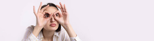 Close up portrait of attractive quirky young woman making binoculars with hands showing ok gesture on white studio background.