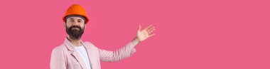 Genç inşaat mühendisinin portresi kırmızı stüdyo arka planında duran pembe ceketli turuncu bir şapka giyer. Adam eliyle işaret ediyor..