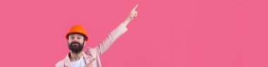 Genç inşaat mühendisinin portresi kırmızı stüdyo arka planında duran pembe ceketli turuncu bir şapka giyer. Adam eliyle işaret ediyor..