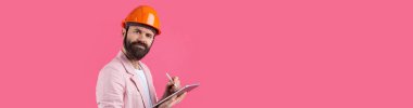 Genç inşaat mühendisinin portresi kırmızı stüdyo arka planında duran pembe ceketli turuncu bir şapka giyer. Elinde tablet olan bir adam..