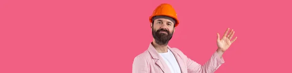 若い建設エンジニアの肖像画は 赤いスタジオの背景に立ってピンクのジャケットで オレンジのハード帽子を着用してください 男は手で指差す — ストック写真