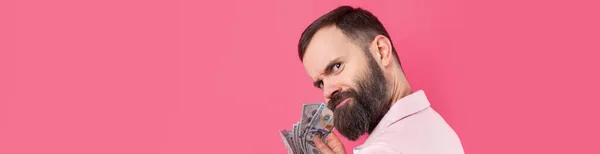 一个心满意足的年轻商人的画像 他身穿粉色夹克 留着胡子 向我们展示在红色工作室背景下的美元钞票 尝起来像钱的味道 — 图库照片