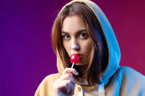 Portrait of a woman in a hoodie, girls licking a red round lollipop with beautiful makeup on a purple trendy background in the studio.