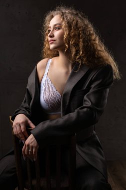 Stüdyoda uzun saçlı, ceketli, kızıl saçlı, kıvırcık bir kızın portresi. Kıvrımlı göğüslerini sütyen içinde gösteriyor..