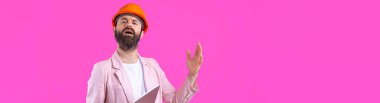 Genç inşaat mühendisinin portresi kırmızı stüdyo arka planında duran pembe ceketli turuncu bir şapka giyer. Elinde tablet olan bir adam..