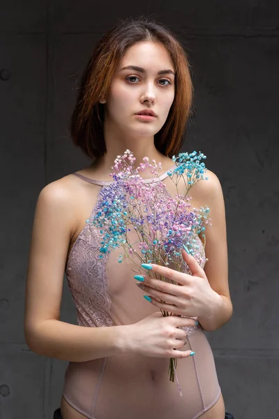 A beautiful sexy lady in an elegant nude bodysuit holds flowers in her hands. Fashion beauty portrait of fashion model girl in studio.