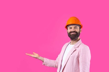 Genç inşaat mühendisinin portresi kırmızı stüdyo arka planında duran pembe ceketli turuncu bir şapka giyer. Adam eliyle işaret ediyor..