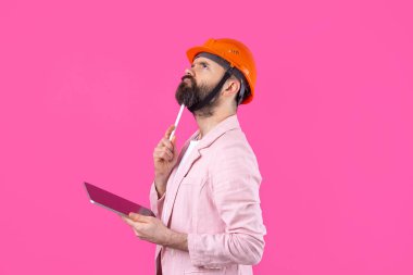 Genç inşaat mühendisinin portresi kırmızı stüdyo arka planında duran pembe ceketli turuncu bir şapka giyer. Elinde tablet olan bir adam..