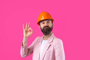 Genç inşaat mühendisinin portresi kırmızı stüdyo arka planında duran pembe ceketli turuncu bir şapka giyer. Adam eliyle işaret ediyor..