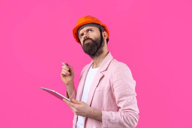 Genç inşaat mühendisinin portresi kırmızı stüdyo arka planında duran pembe ceketli turuncu bir şapka giyer. Elinde tablet olan bir adam..