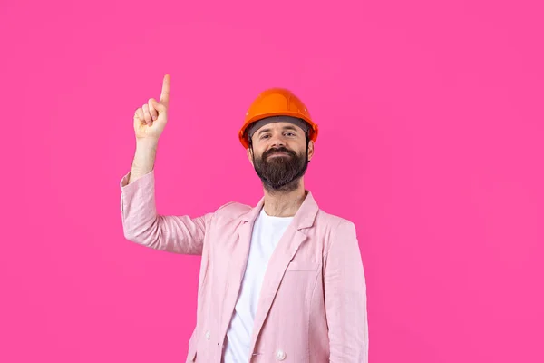 Portrait of young construction engineer wear orange hard hat, in a pink jacket standing on red studio background. The man points with his hand.
