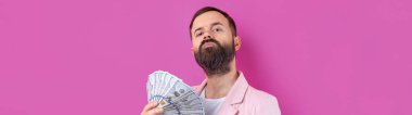Pembe ceketli sakallı, mutlu genç bir iş adamının portresi kırmızı bir stüdyonun arka planında bize dolar banknotları gösteriyor. Tat, para kokusu..